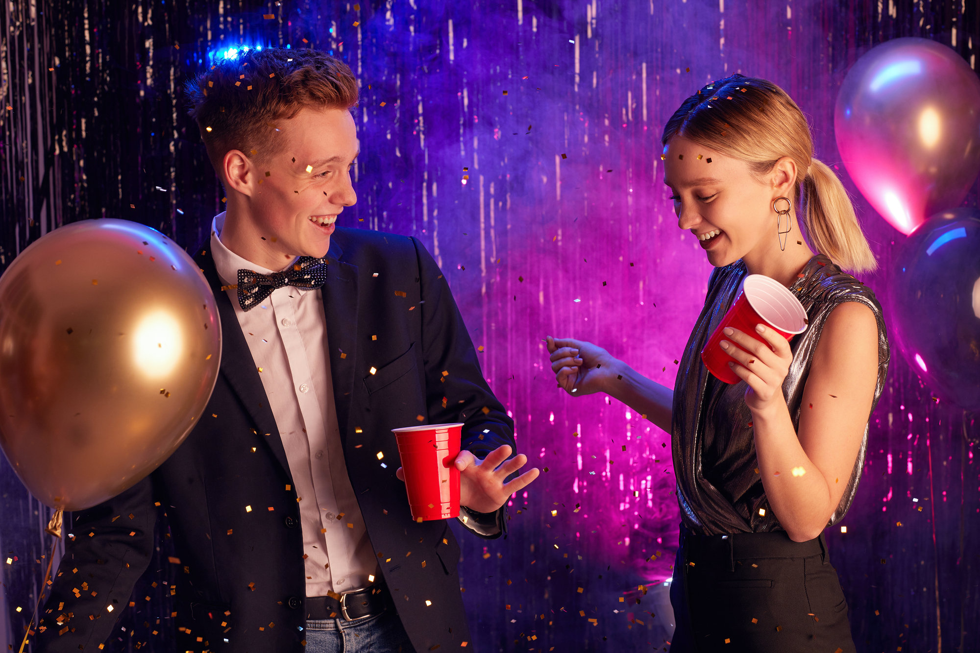 Waist up portrait of happy teenage couple dancing in decorated hall and holding red cups while enjoying prom night or party