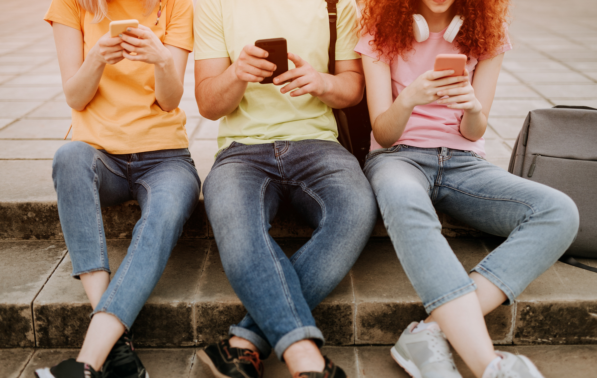 Group of crop students in casual clothes sitting together on stairs on city street and using mobile phones checking messages and browsing internet